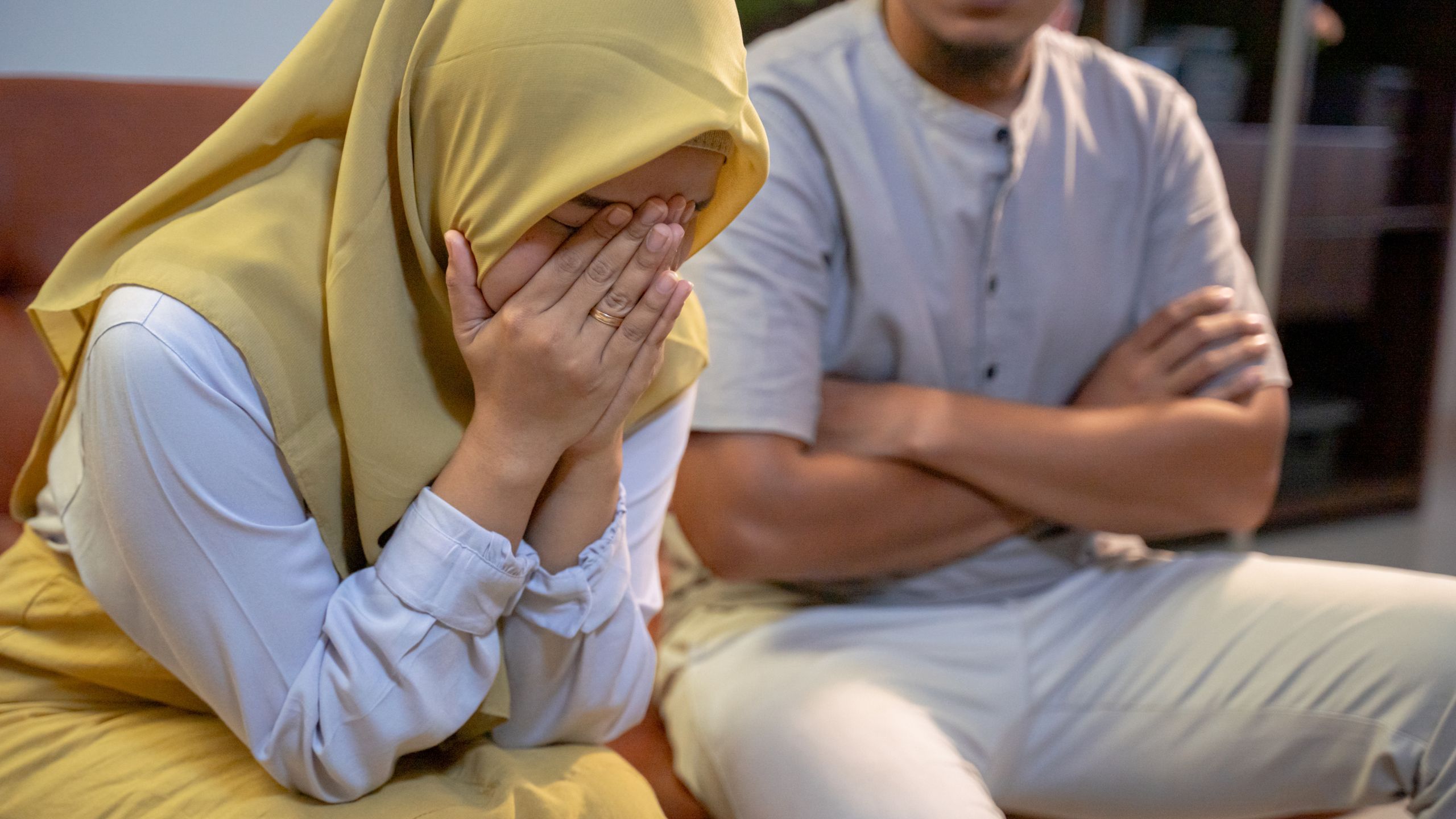 Wife sitting down crying with her hands convering her face and her husband is sitting right beside her with his hands folded