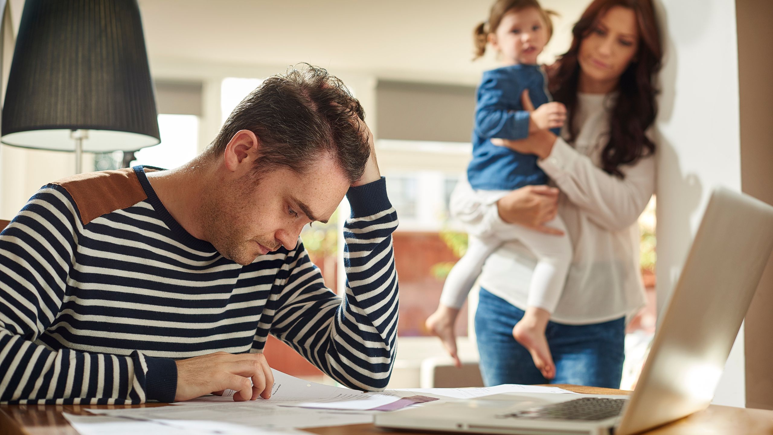 Mother carrying daughter while standing up and father sitting on a table working on a laptop - Family Emotional Quotes in Telugu